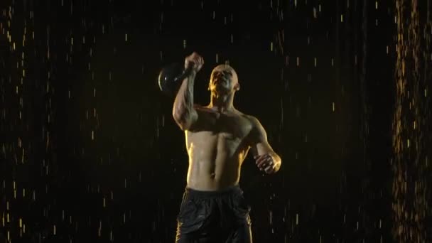 Halterofilista levanta o kettlebell acima de sua cabeça. Treino de força. Forte atleta crossfit. Gotas de chuva caem sobre o corpo muscular molhado. Fundo preto com luz. Movimento lento. Fechar. — Vídeo de Stock