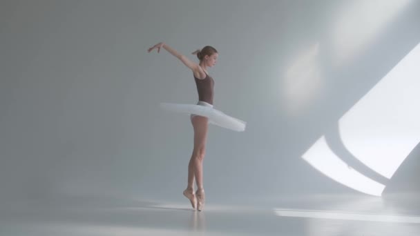 Профессиональная балерина в белой пачке танцует в большом тренировочном зале. Девушка делает танцевальные шаги в костюме сцены. Съемка на белом фоне в просторной и ярко освещенной студии. Медленное движение. — стоковое видео