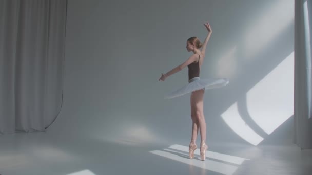 La bailarina hace ejercicios de ballet en vestido de escenario con la espalda abierta. Ensayos de danza se mueve en el amplio estudio bañado por la luz del sol. Movimiento lento. — Vídeos de Stock