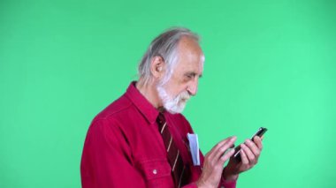 70 'li yaşlarda mutlu yaşlı bir adamın portresi yeşil arka planda izole edilmiş telefonuyla mesajlaşan biriyle iletişim kuruyor..