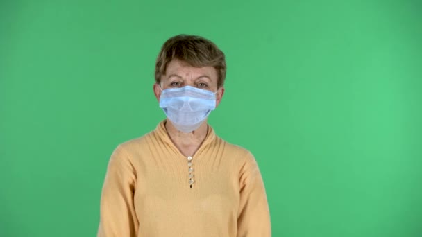 Portrait of elderly woman puts a medical mask on her face to protecting yourself from coronavirus pandemic isolated over green background. — Stock Video