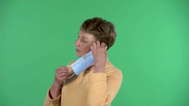 Portrait of elderly woman puts a medical mask on her face to protecting yourself from coronavirus pandemic isolated over green background. Profile view — Stock Video