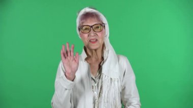 Yaşlı stil sahibi kadının portresi el sallıyor ve el sallıyor. Yeşil arka planda izole bir şekilde buraya geliyor..