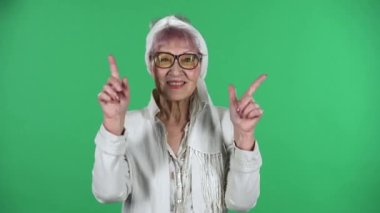 Yaşlı stil sahibi kadının portresi gülümsüyor ve kalbi parmaklarıyla gösteriyor sonra yeşil arka planda izole bir şekilde öpüşüyor..