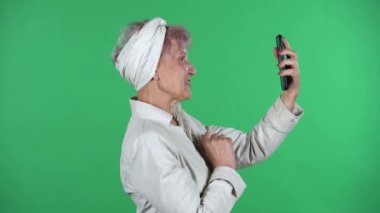 Yaşlı stil sahibi kadın yeşil arka planda izole edilmiş cep telefonu kullanarak görüntülü konuşma yapıyor. Profil