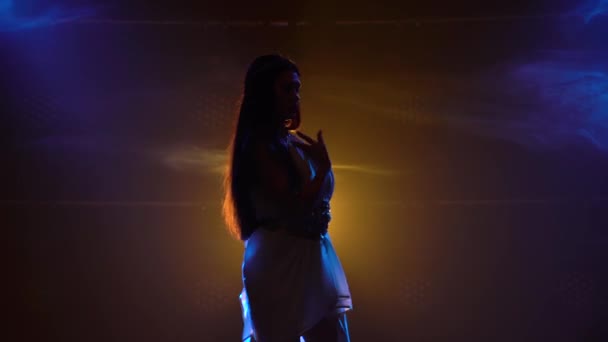 Brandende brunette danst in een nachtclub in het donker en dynamische neonlicht. Een rokerige achtergrond. Dansen maar. Silhouet meisje in grieks grieks godinnen jurk en krans high fashion. — Stockvideo