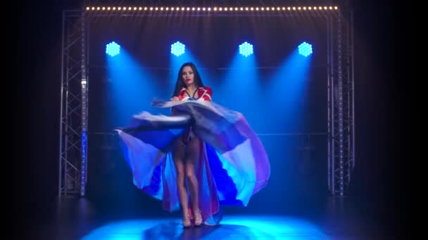 Smal high girl dansar i en sexig kostym i färgen av den engelska flaggan och strass. Attraktiv brunett viftar med kjolen i en mörk studio med rök och blått neonljus. — Stockvideo