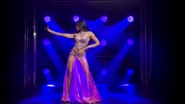 Doğulu dansçı dans ediyor ve vücudunun esnekliğini ve çekiciliğini gösteriyor. Mavi arka plan neon ışıkları. Takma elmaslarla süslenmiş sosyetik kostümlü genç bir kız..