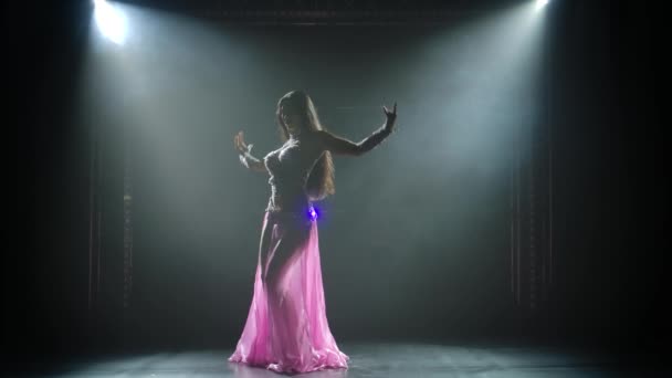 Esbelta bailarina oriental alta está bailando sobre un fondo negro en el humo. Silueta de una joven con un disfraz oriental salpicado de pedrería. Movimiento lento. — Vídeo de stock