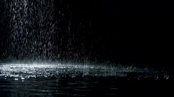 A mennyezetről fröccsenő eső esik a vízfelszínre, amely világít. Fekete háttér. Fehér neon világítás a sötét stúdióban. Lassú mozgás. Közelről.. — Stock videók