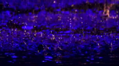 Büyük yağmur damlaları, mavi bir yağmurla aydınlanan suyun yüzeyinde küçük sıçramalar yaratır. Siyah arka planda çekilmiş. Kapatın. Yavaş çekim.