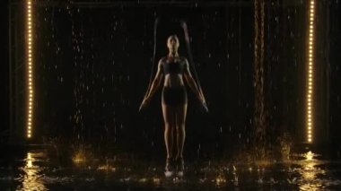 Genç kaslı kadın atlama halatıyla kardiyo çalışması yapıyor. Yağmurda stüdyoda Crossfit idmanı. Sprey kadınların farklı yönlerine sıçrıyor. Yavaş çekim.