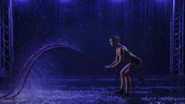 Atletik genç kadın yağmur altında savaş halatıyla çapraz egzersizler yapıyor. Siyah arka plan. Yavaş çekim.