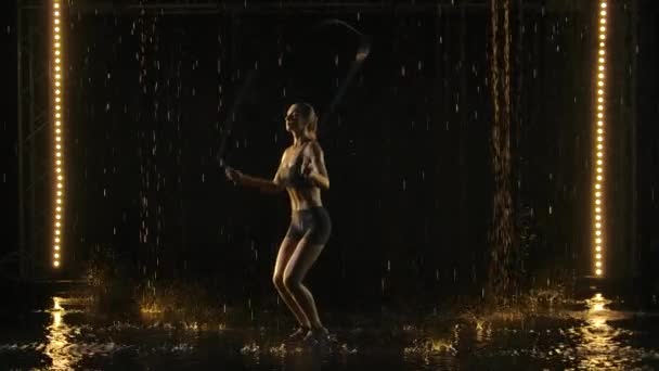 Fiatal, felnőtt, sportos nő ugrókötél edzést végez sstudiókban az esőben. A kötél megüti a víz felszínét, és sok fröccsenést okoz. Fekete háttér fénnyel. Lassú mozgás.. — Stock videók