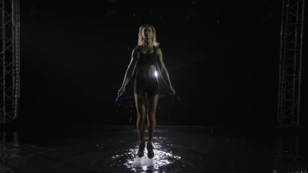 Mujer deportiva en ropa deportiva negra haciendo ejercicios de salto con una cuerda de salto. Entrenamiento Crossfit en el estudio bajo la lluvia. El spray se dispersa en diferentes direcciones de los saltos de las mujeres. Movimiento lento. — Vídeos de Stock