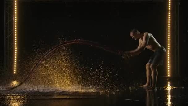 Masculine sportsman doing workout using a battle rope. Side view. The man slams the battle ropes hard on the surface of the water, splashing it. Black background and stage light. Slow motion. — Stock Video