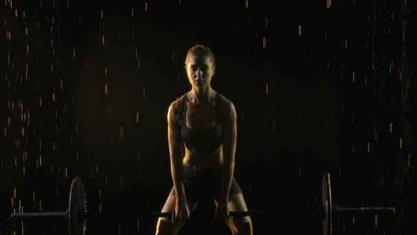 Retrato de hembra realizando ejercicio de levantamiento de pesas con barra de peso. Mujer joven de confianza haciendo levantamiento de pesas barra de entrenamiento. Filmado en un estudio oscuro bajo la lluvia y la luz puesta en escena. En cámara lenta. De cerca.. — Vídeos de Stock