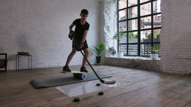 Een leuke jongen hockey speler staat op een balance board en traint met een stok en puck op een gladde ondergrond binnen. Langzame beweging. — Stockvideo