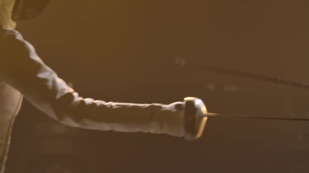 Руки фехтовальщика в белом костюме держат меч и соревнуются с неизвестным противником. Концепция спортивного фехтования. Снимок сделан на черном фоне с мягким желтым светом. Медленное движение. Закрыть. — стоковое видео