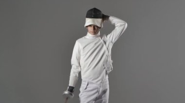 Maske takan, beyaz eskrim kıyafeti giyen ve önünde kılıç tutan genç bir adamın portresi. Gri arka planda izole edilmiş. Ağır çekim. Kapat..