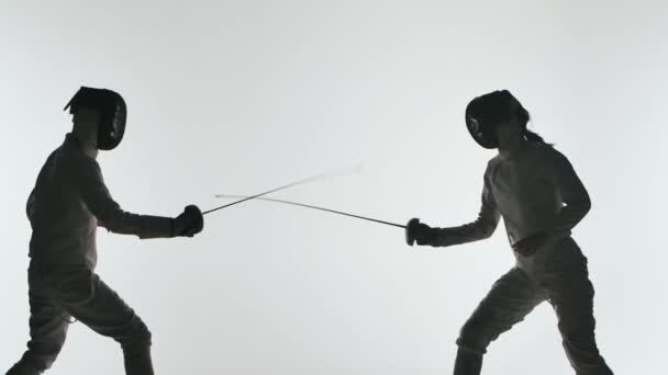 Donkere silhouetten Twee atleten met helmen en witte uniformen tonen meesterlijk zwaardvechten. Ze ontwijken, springen en duwen en springen. Geschoten op witte achtergrond. Langzame beweging. — Stockvideo