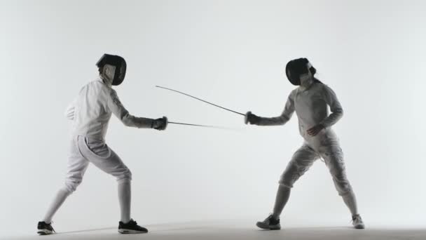 Zwei Athleten mit Helmen und weißen Uniformen zeigen in ihrem Florettkampf meisterliche Schwertkunst. Sie weichen aus, springen und stoßen und fallen aus. Schuss isoliert auf weißem Hintergrund. Zeitlupe. — Stockvideo