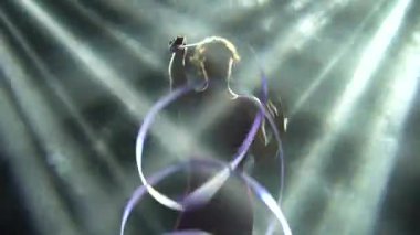 Dumanlı bir arka planda, genç bir sporcu kadın aktif olarak spot ışıklarında ipek bir kurdele ile antrenman yapıyor. Esnek, ince bir vücudun siluetleri. Aktif yaşam tarzı konsepti. Kapat..