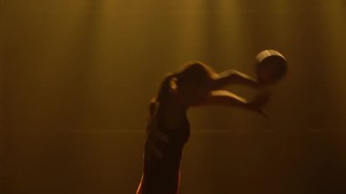 Genç bir jimnastikçi bir topla egzersiz yapıyor. El ve vücut sallıyor ve takla atıyor. Stüdyoda, üzerinde duman ve sarı yumuşak ışık olan bir kadının silueti. Kapat..