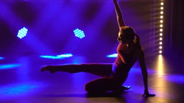 Genç ritmik jimnastikçi akrobatik hareketler sergiliyor. Stüdyoda, arka plandaki mor spot ışıklarına karşı spor eğitmeni. Kapat..