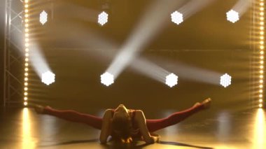 Sıkı spor bir vücut giysisi giymiş bir jimnastikçi kız bölünmüş bir şekilde oturur ve harika bir esneme hareketi gösterir. Stüdyoda sigara dumanı ve sarı ışıkla profesyonel sporcu kadın eğitimi. Kapat..