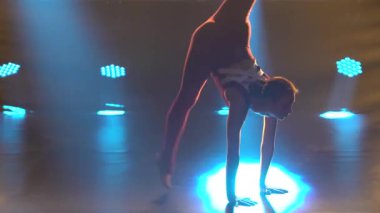 Sıkı spor bir vücut giysisi giymiş bir jimnastikçi kız bölünmüş bir şekilde oturur ve harika bir esneme hareketi gösterir. Stüdyoda duman ve hafif ışıkla profesyonel sporcu kadın eğitimi. Kapat..