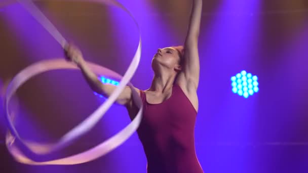 Rytmisk gymnastik. Ung atletisk kvinna utför en övning med ett band. Skjuten i en mörk studio med lila neonljus i bakgrunden. Närbild. — Stockvideo
