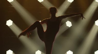 Genç, esnek bir bayan ritmik jimnastikçi tarafından yapılan akrobatik egzersizler. Spot ışığında ince bir vücudun koyu bir silueti. Kapat..