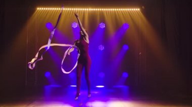 Ritmik jimnastik. Genç atletik kadın bir kasetle egzersiz yapıyor. Arka planda mor neon ışıkları olan karanlık bir stüdyoda çekilmiş. Yavaş çekim.