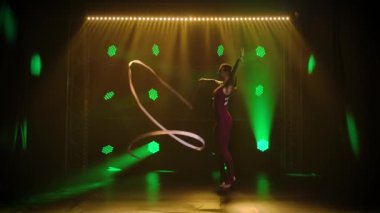 Genç sporcu kız jimnastikçi vücudunun esnekliğini göstermek için kurdeleyle akrobatik egzersizler yapıyor. Yeşil ışıklı karanlık bir stüdyoda çekilmiş. Yavaş çekim.