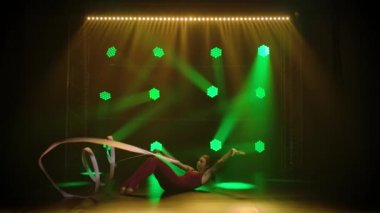 Genç sporcu kız jimnastikçi vücudunun esnekliğini göstermek için kurdeleyle akrobatik egzersizler yapıyor. Yeşil ışıklı karanlık bir stüdyoda çekilmiş. Yavaş çekim.