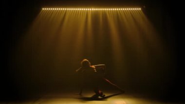 Genç bir jimnastikçi bir topla egzersiz yapıyor. El ve vücut sallıyor ve takla atıyor. Stüdyoda, üzerinde duman ve sarı yumuşak ışık olan bir kadının silueti. Yavaş çekim.