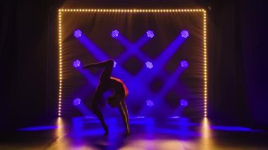 Genç ritmik jimnastikçi akrobatik hareketler sergiliyor. Stüdyoda, arka plandaki mor spot ışıklarına karşı spor eğitmeni. Yavaş çekim.