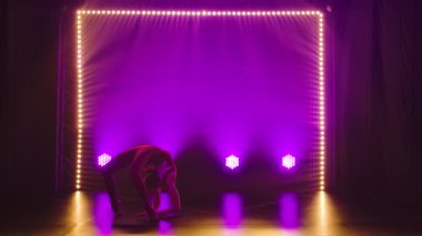 Esnek profesyonel bir bayan atlet, ritmik jimnastiğin karmaşık akrobatik unsurlarını sergiler. Neon ışıkları olan karanlık bir stüdyoda çekilmiş. Siluet. Yavaş çekim.