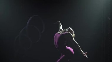 Cılız, genç bir kadın jimnastikçinin silueti. Sıkı bir vücut, kurdeleyle jimnastik yapıyor. Spot ışığında siyah dumanlı bir arka plan. Ağır çekim. Kapat..