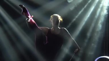 Dumanlı bir arka planda, genç bir sporcu kadın aktif olarak spot ışıklarında ipek bir kurdele ile antrenman yapıyor. Esnek, ince bir vücudun siluetleri. Aktif yaşam tarzı konsepti. Ağır çekim. Kapat..