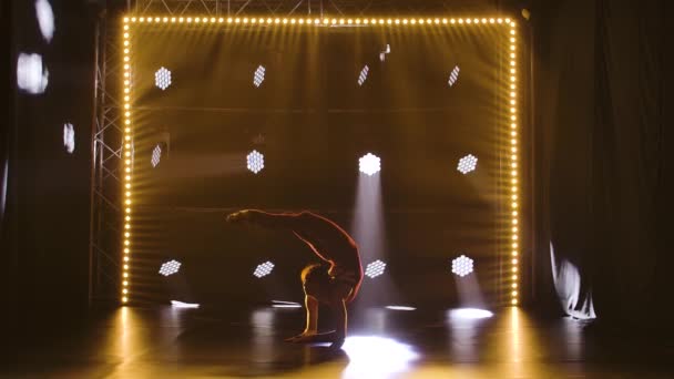 Une athlète féminine professionnelle flexible exécute des éléments acrobatiques complexes de gymnastique rythmique. Tourné dans un studio sombre avec des néons. Silhouette. Mouvement lent. — Video