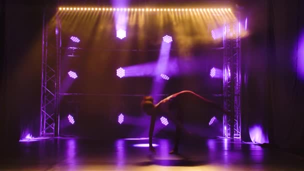 Une athlète féminine professionnelle flexible exécute des éléments acrobatiques complexes de gymnastique rythmique. Tourné dans un studio sombre avec des néons. Silhouette. Mouvement lent. — Video