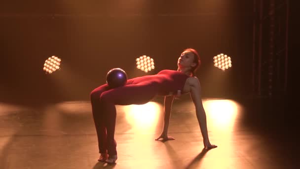 Sportovní dívka gymnastka provádí akrobatické cvičení s míčem a dělá protahování, ukazuje pružnost svého těla. Zastřelen v tmavém studiu s neonovými světly. Zpomal. Zavřít. — Stock video
