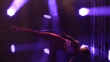 İnce esnek profesyonel bayan atlet ritmik jimnastiğin karmaşık akrobatik unsurlarını sergiliyor. Mor neon ışıkları olan karanlık bir stüdyoda çekilmiş. Siluet. Kapatın. Yavaş çekim.