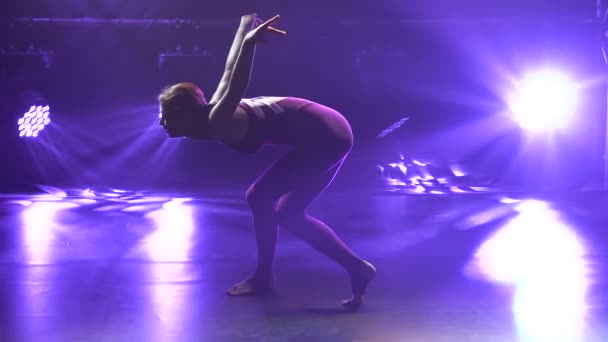 Slim athlète féminine professionnelle flexible effectue des éléments acrobatiques complexes de la gymnastique rythmique. Tourné dans un studio sombre avec des néons violets. Silhouette. Ferme là. Mouvement lent. — Video