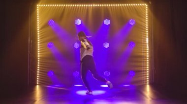 Spor kıyafetleri giymiş, break dans tarzında gösteri yapan genç aktif bir adam. Dans stüdyosunda eğitim. Koyu dumanlı arka plan ve mavi neon ışıklar. Yavaş çekim.