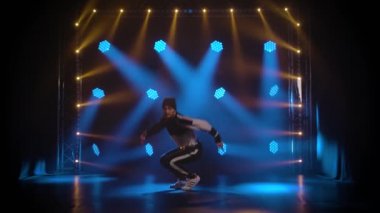 Sokak tarzında zıplayan ve dans eden genç biri. Stüdyoda eşofman giyen ve spor ayakkabı eğitimi alan bir adam. Mavi ışıklı siyah arka plan. Yavaş çekim.