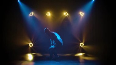 Stüdyo ışıklandırmasının arka planında break dans yapan genç sporcu. Eşofmanlı siluet dansçı adam karmaşık hareketler sergiliyor. Hayat tarzı olarak sokak dansı. Yavaş çekim.