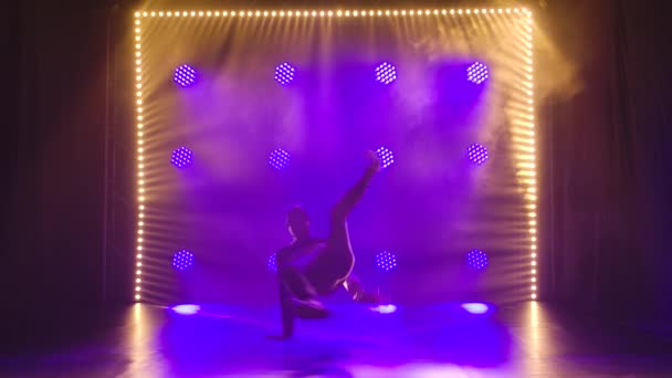 Break dance framförd av en ung kille i träningsoverall. Silhuett av en manlig dansare som snurrar på golvet och utför grundläggande rörelser. Mörk studio med ljus. Långsamma rörelser. — Stockvideo